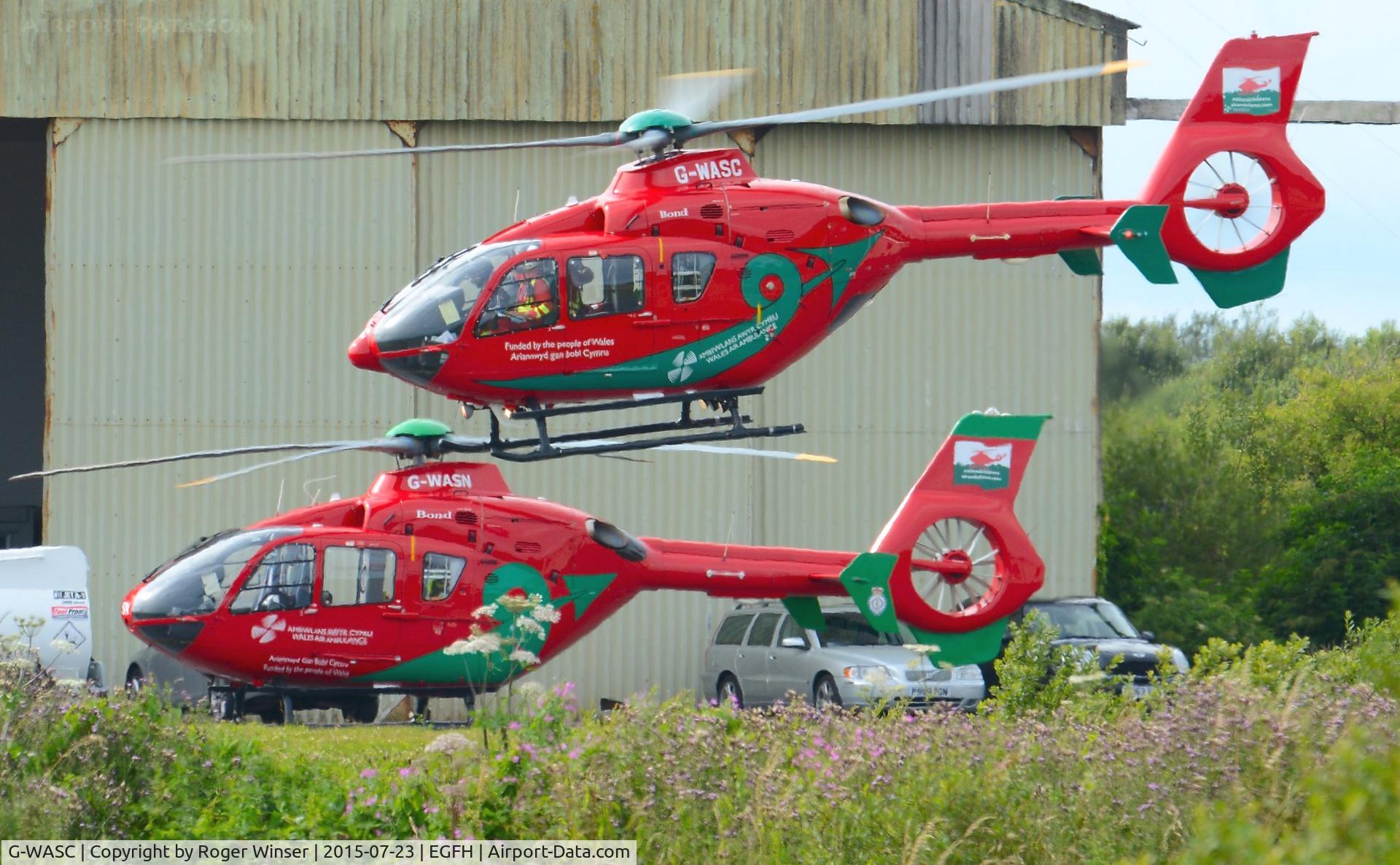 G-WASC, 2012 Eurocopter EC-135T-2+ C/N 1074, Visiting Wales Air Ambulance helicopter (Helimed 59) departing the South Wales Air Ambulance base with resident air ambulance helicopter (G-WASN Helimed 57) on the apron.