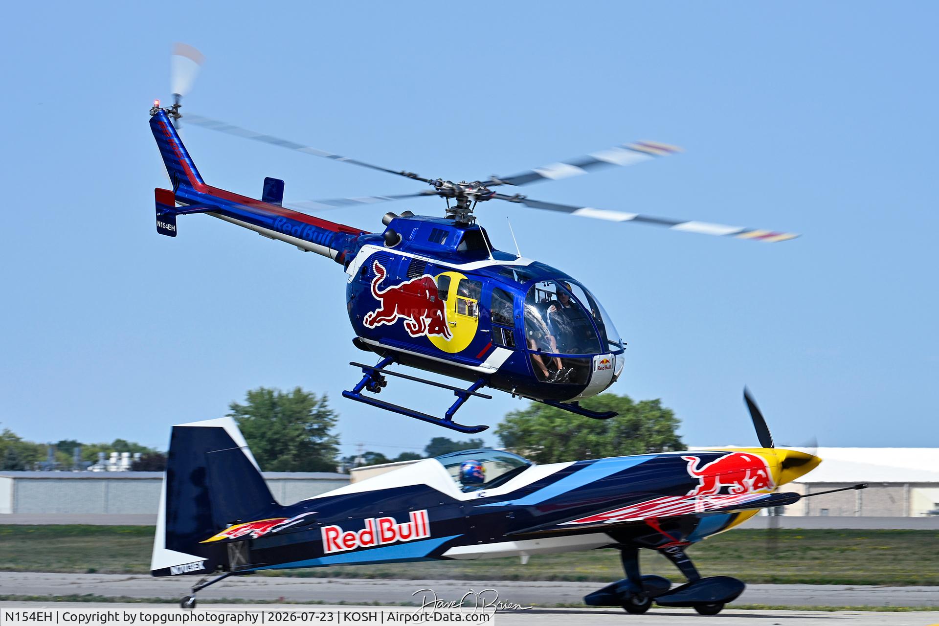 Aircraft N154EH, 1985 MBB Bo-105CBS-4, c/n S-704, Red Bull helo taking off while the Red Bull Extra holds on the taxiway, at KOSH, photo by topgunphotography